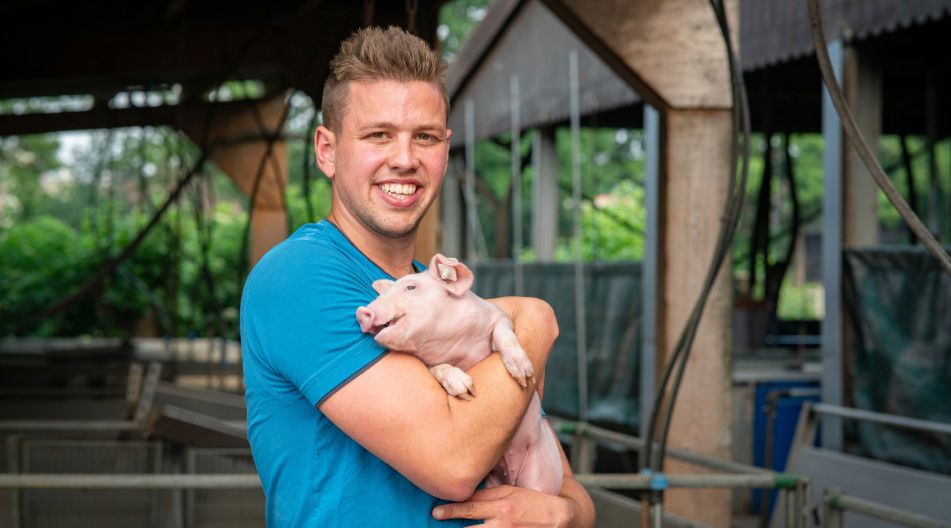 Ein junger Bauer hält vor einem Stall lächelnd ein Schwein im Arm. Ein junger Bauer hält vor einem Stall lächelnd ein Schwein im Arm.
