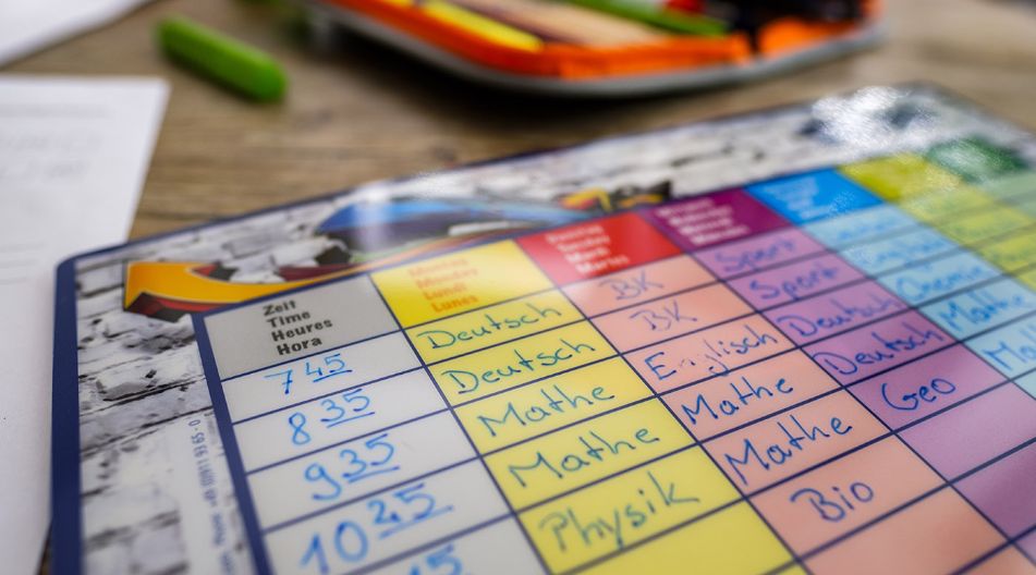 Ein Stundenplan mit handschriftlich eingetragenen Fächern wie Deutsch, Mathe und Englisch liegt auf einem Tisch. Im Hintergrund sind unscharf Stifte und weitere Schulutensilien zu erkennen. Ein Stundenplan mit handschriftlich eingetragenen Fächern wie Deutsch, Mathe und Englisch liegt auf einem Tisch. Im Hintergrund sind unscharf Stifte und weitere Schulutensilien zu erkennen.